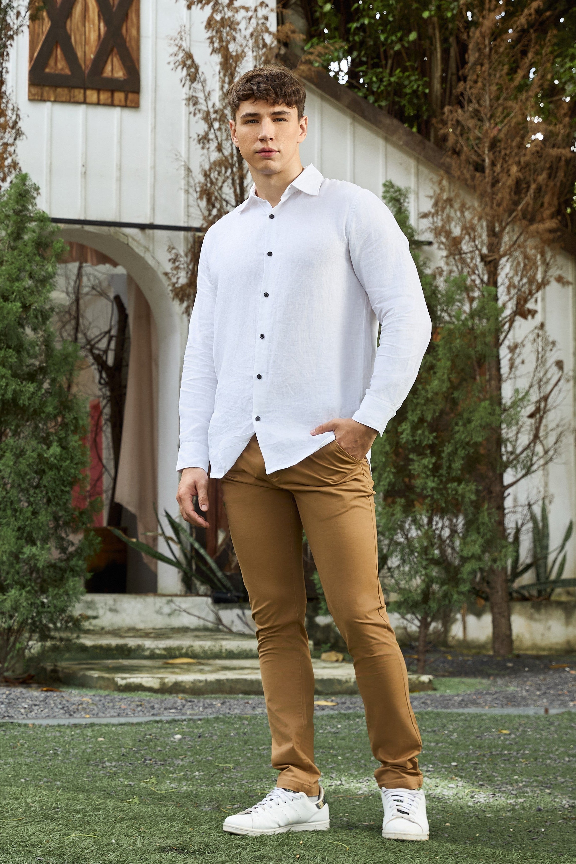 Man wearing a white shirt and brown pants standing outdoors with greenery and a building in the background